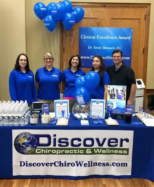 A display table with Discover Chiropractic & Wellness branding, brochures, water bottles, and blue balloons promoting services and awards. Behind the table is Dr. Scott Shepard with another staff members.